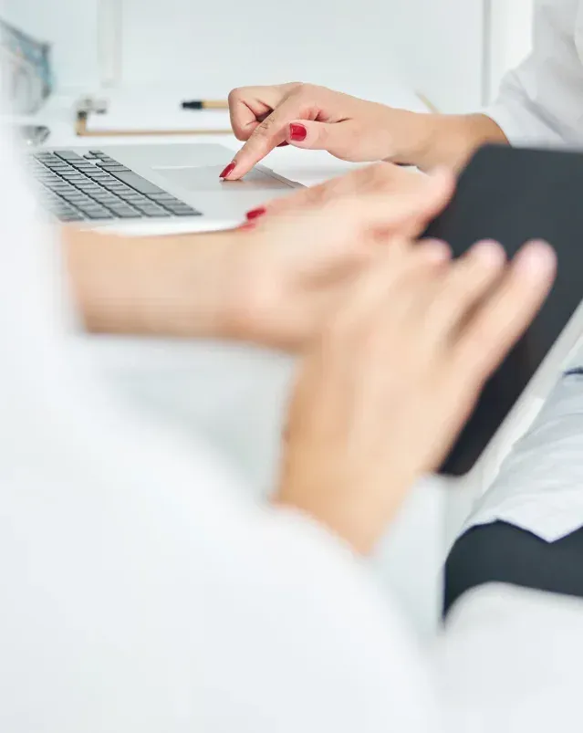 A clinician's hands on a tablet beside a laptop