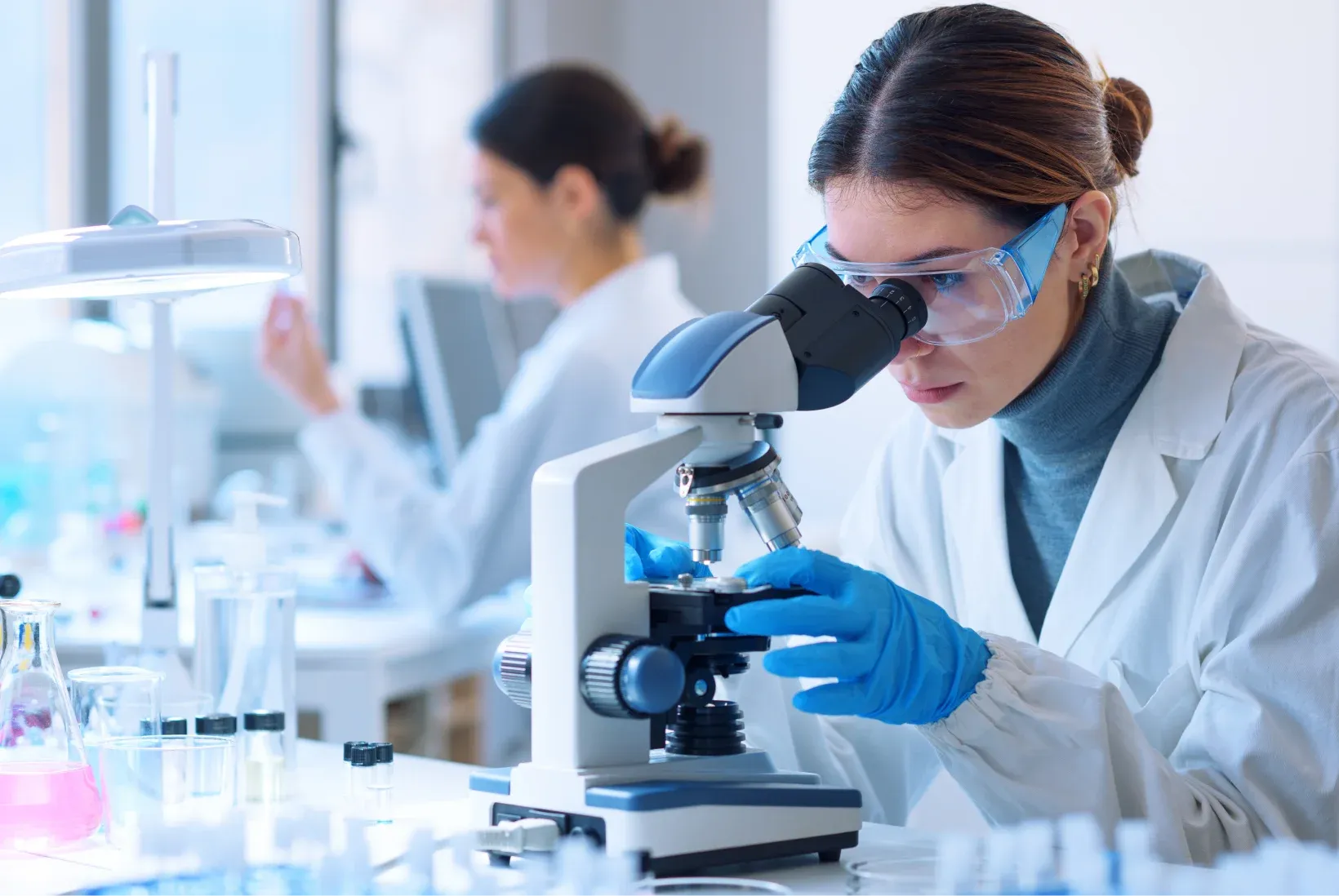 A researcher in a lab coat and safety goggles examining a sample under a microscope, with a colleague working in the background